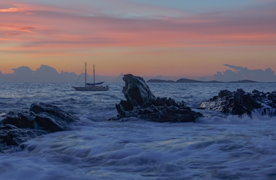 Farbiger Himmel, raues Meer mit Felsen, im Hintergrund ein Schiff.