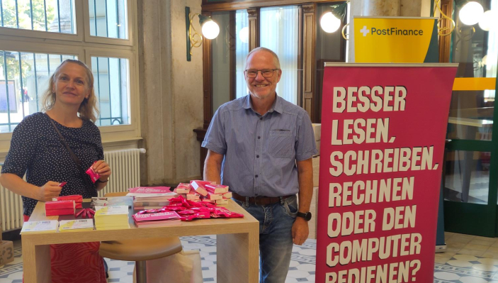 Ein Mann und eine Frau an einem Stand in der Post. Sie machen auf digitale Kompetenzen aufmerksam.
